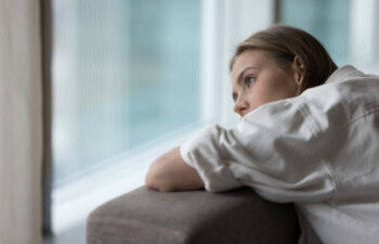 Woman in a white shirt leans on a couch, gazing thoughtfully out a window.